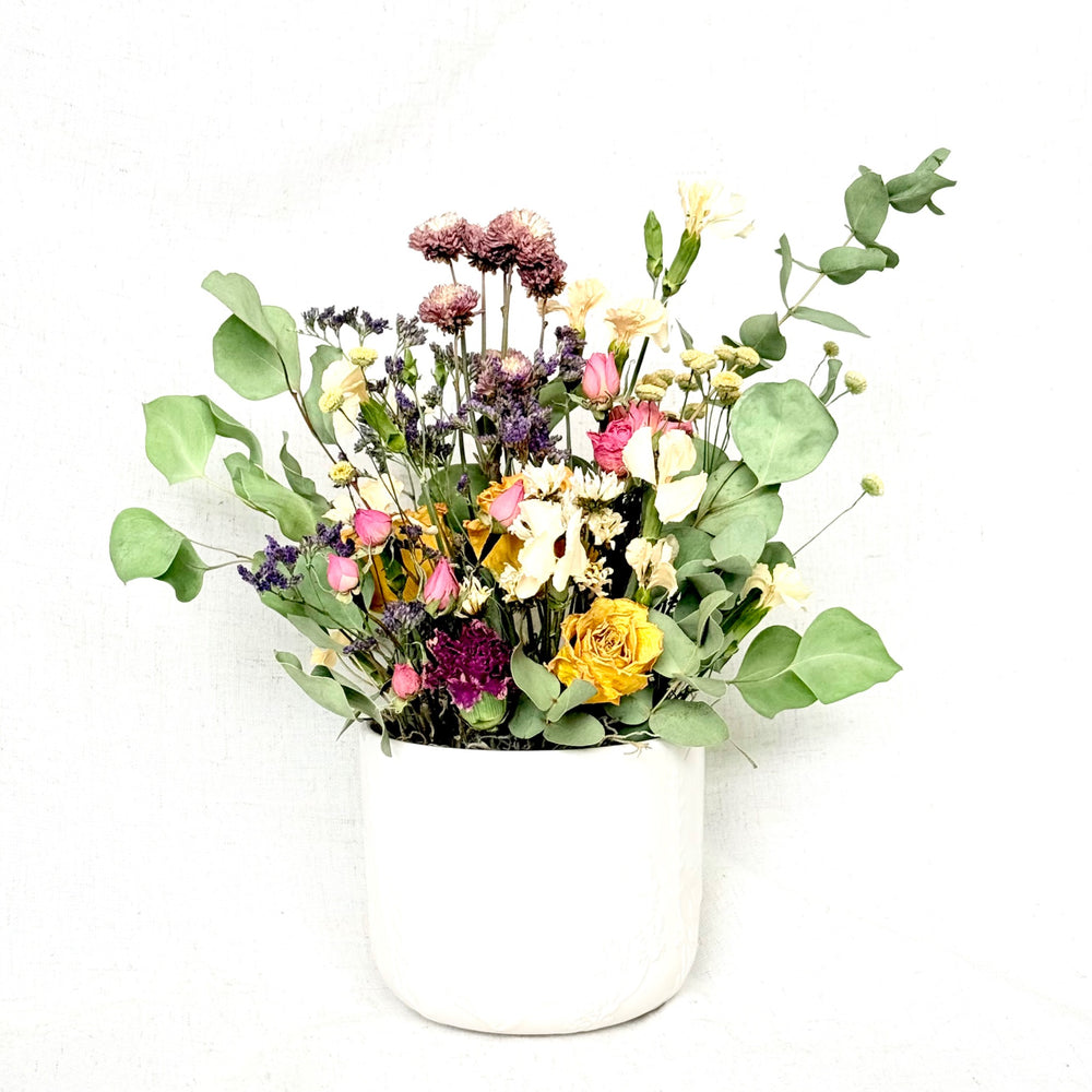 Sunshine Soirée with golden yellow roses, pink buds, purple flowers, and eucalyptus in a white vessel